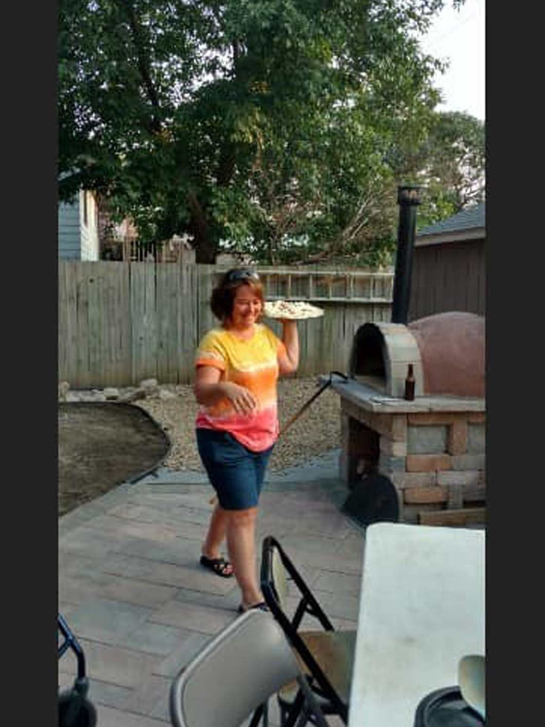 Person in a yellow-orange shirt stands by a brick outdoor pizza oven in a backyard.