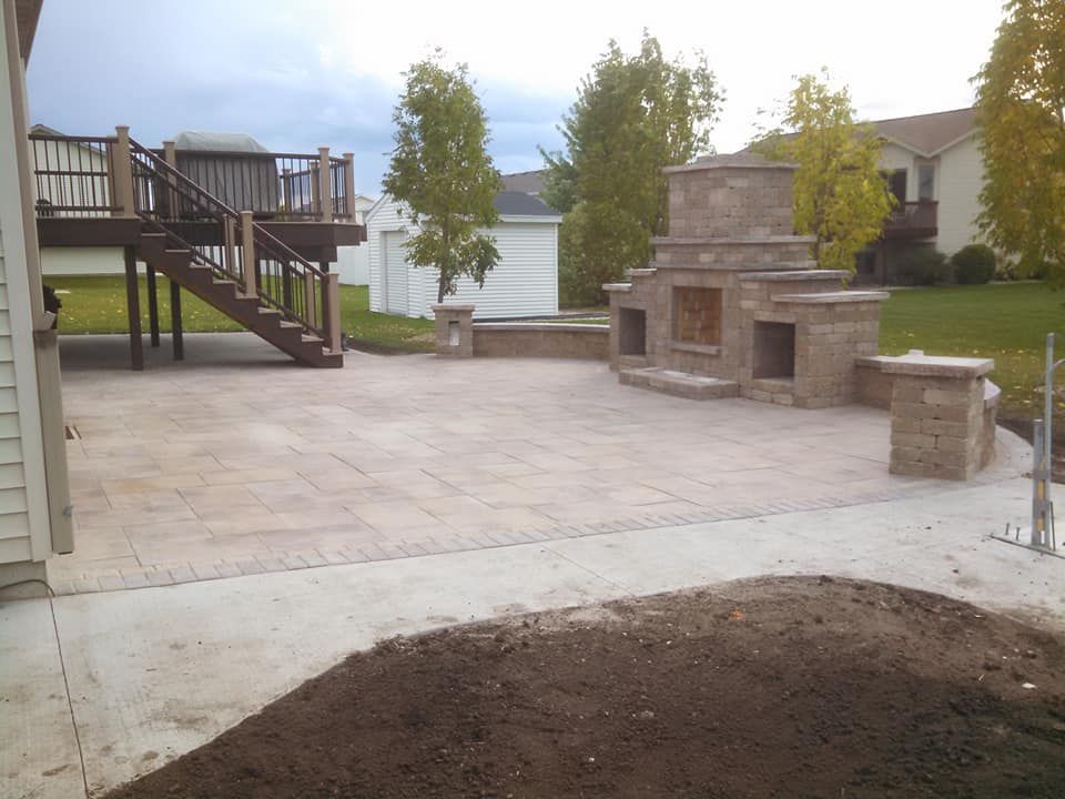 Backyard patio with stone grill, deck stairs, and a dirt pile in the foreground