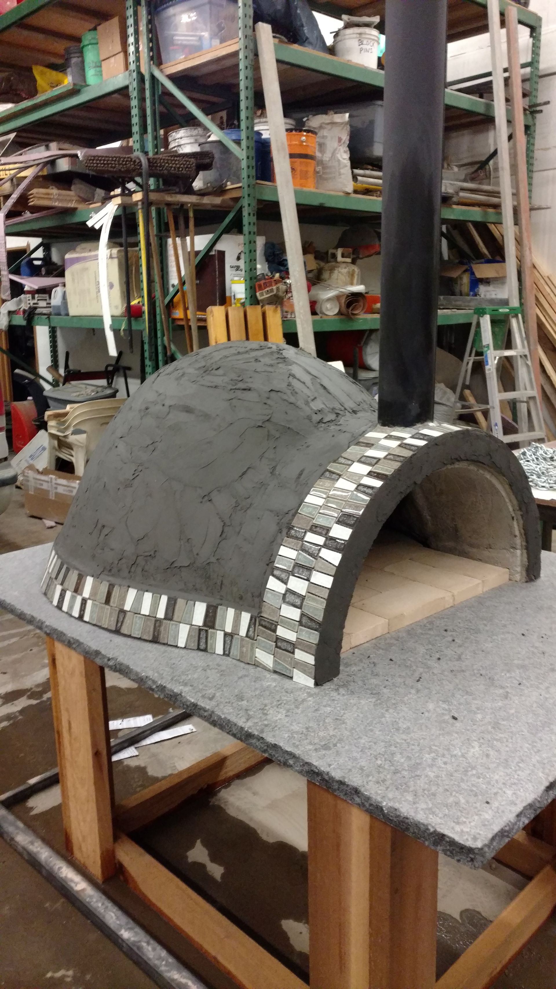 Small dome-shaped oven under construction on a workbench in a workshop, with metal arch framing and gray mortar.