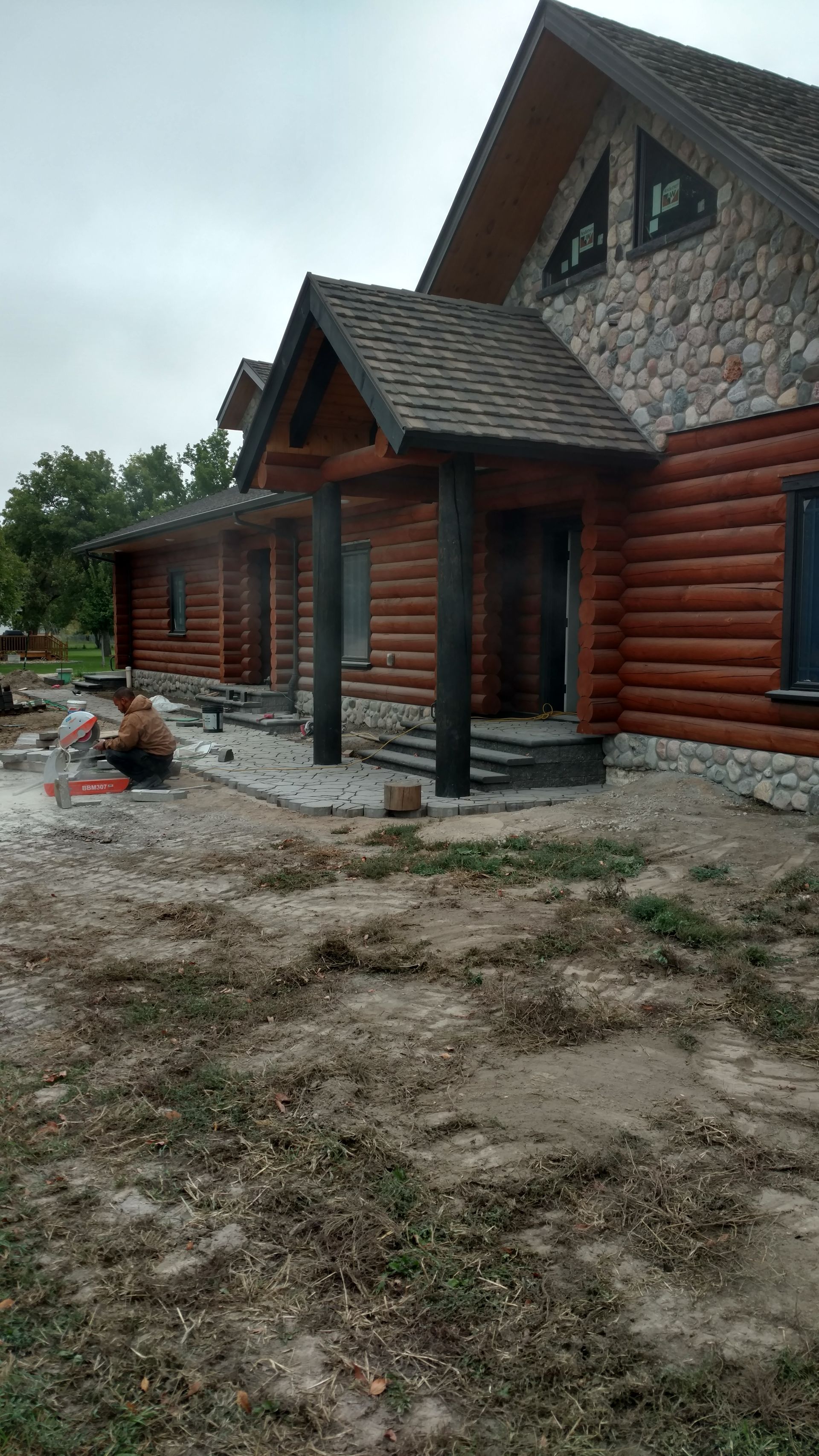 Log cabin-style house under construction with log siding, stone gables, and muddy yard
