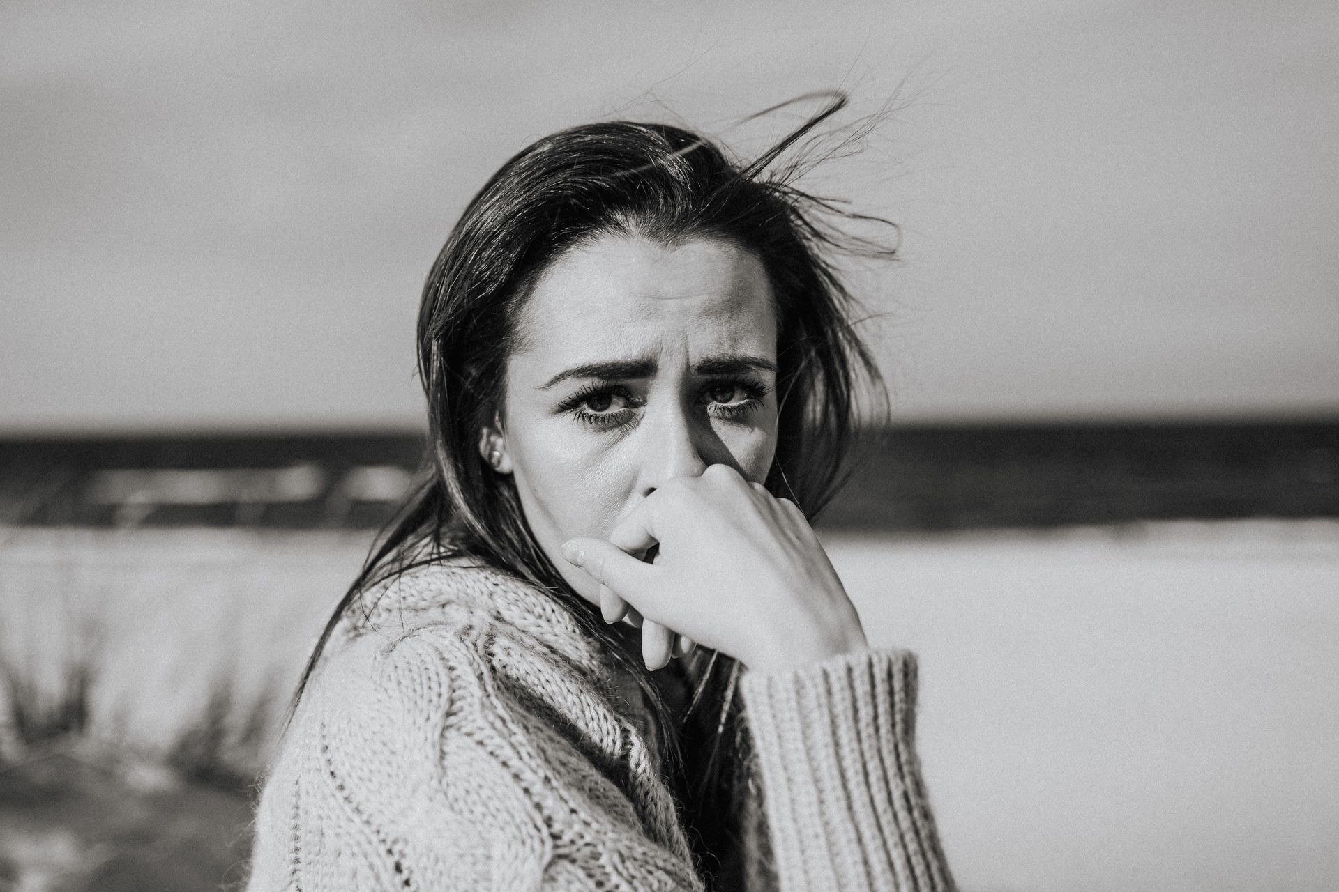 Black-and-white portrait of a person in a sweater, looking toward the camera with a hand near their face outdoors