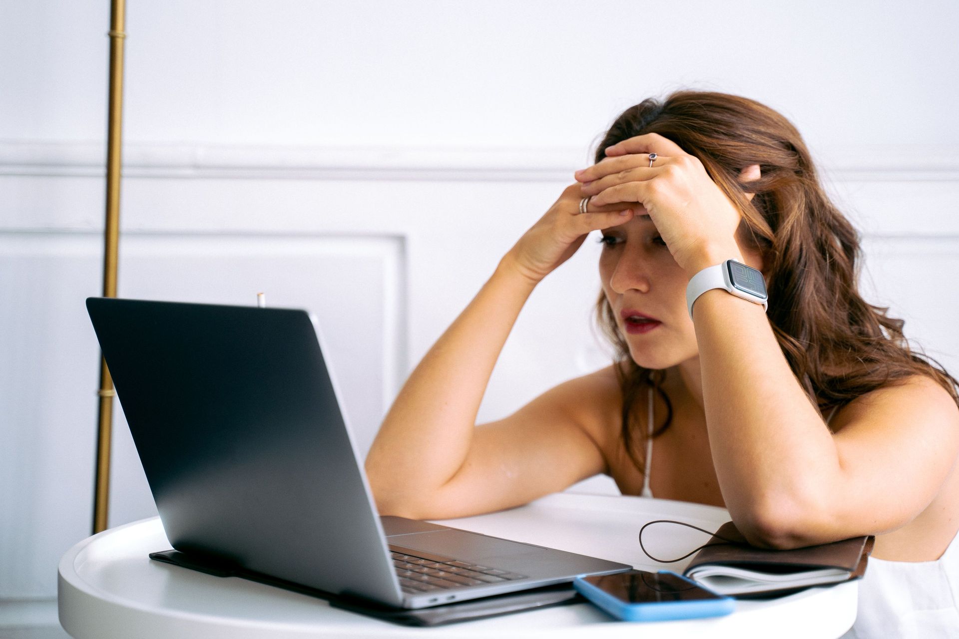 Woman at a laptop, holding her head in frustration at a desk with papers and glasses.