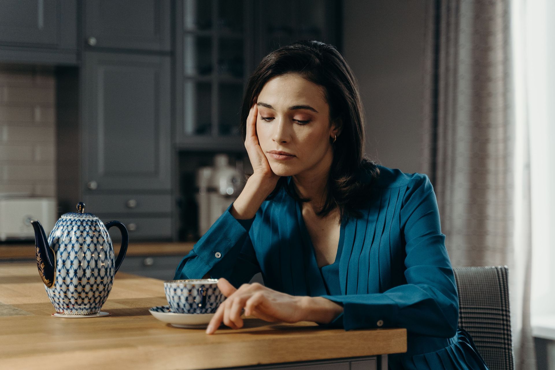 Woman in a teal blouse sits at a table, resting her head beside a cup and kettle in a dim kitchen.