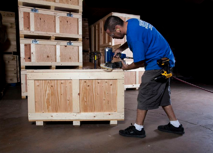 Worker using a blue power drill to assemble a wooden crate in a warehouse