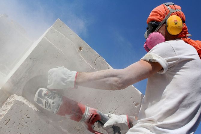 Worker in orange gear cutting a concrete block, with dust cloud and blue sky background