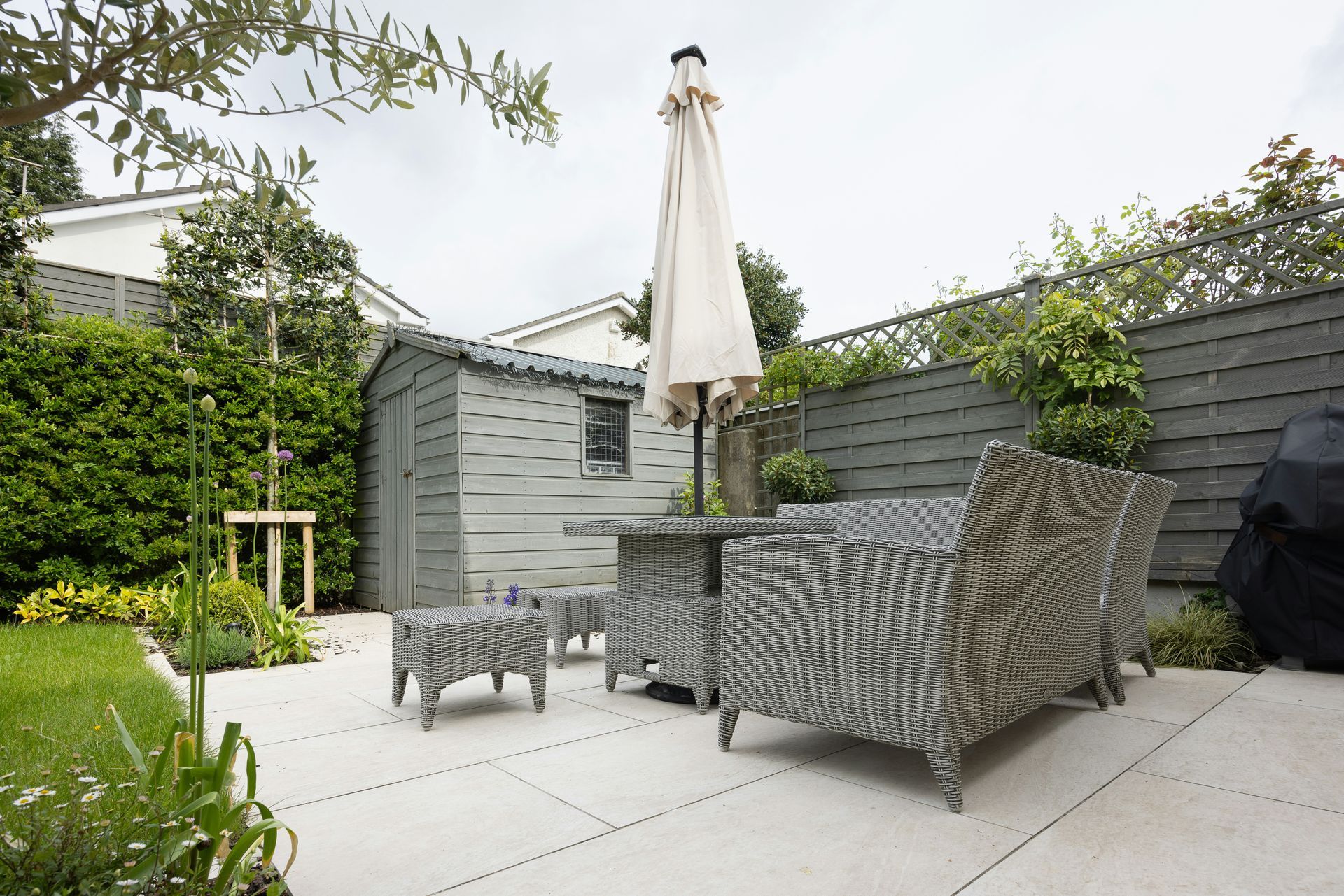 Patio with gray wicker seating, closed umbrella, potted plant, and lawn beside a wooden fence.