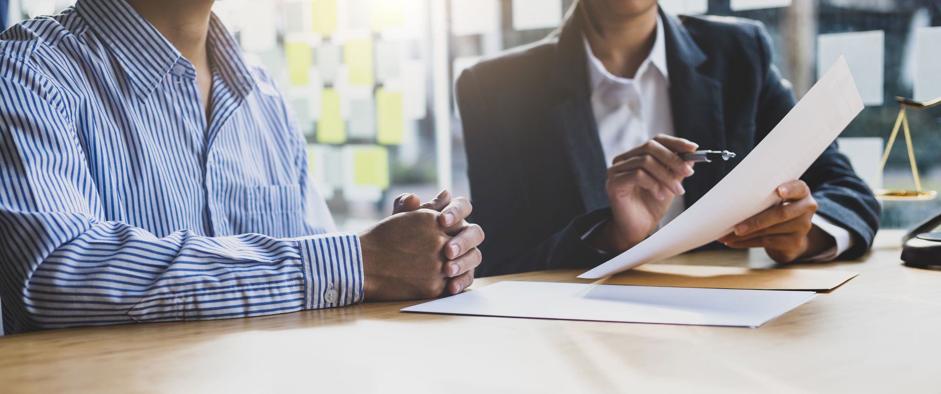 Person in striped shirt clasps hands as a suited advisor reviews paperwork by brass justice scales. Person in striped shirt clasps hands as a suited advisor reviews paperwork by brass justice scales.
