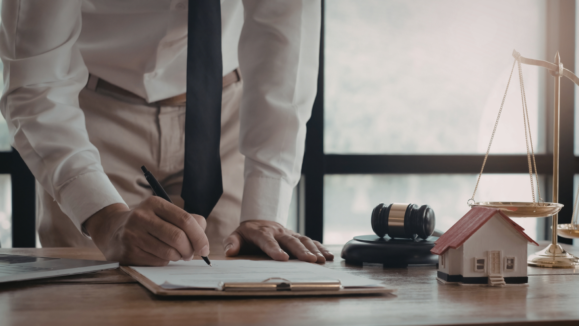 A person in a dress shirt and tie writes on a document next to a small house model and a scale of justice on a desk.