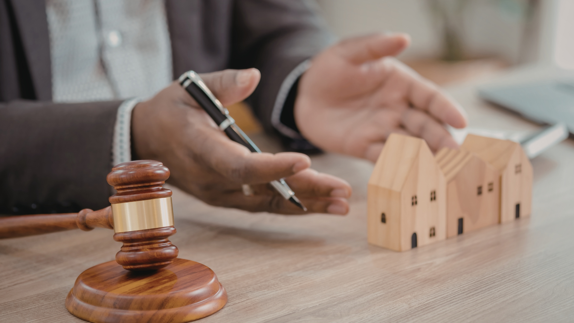 A wooden gavel rests on a desk next to a professional gesturing toward small wooden house models.