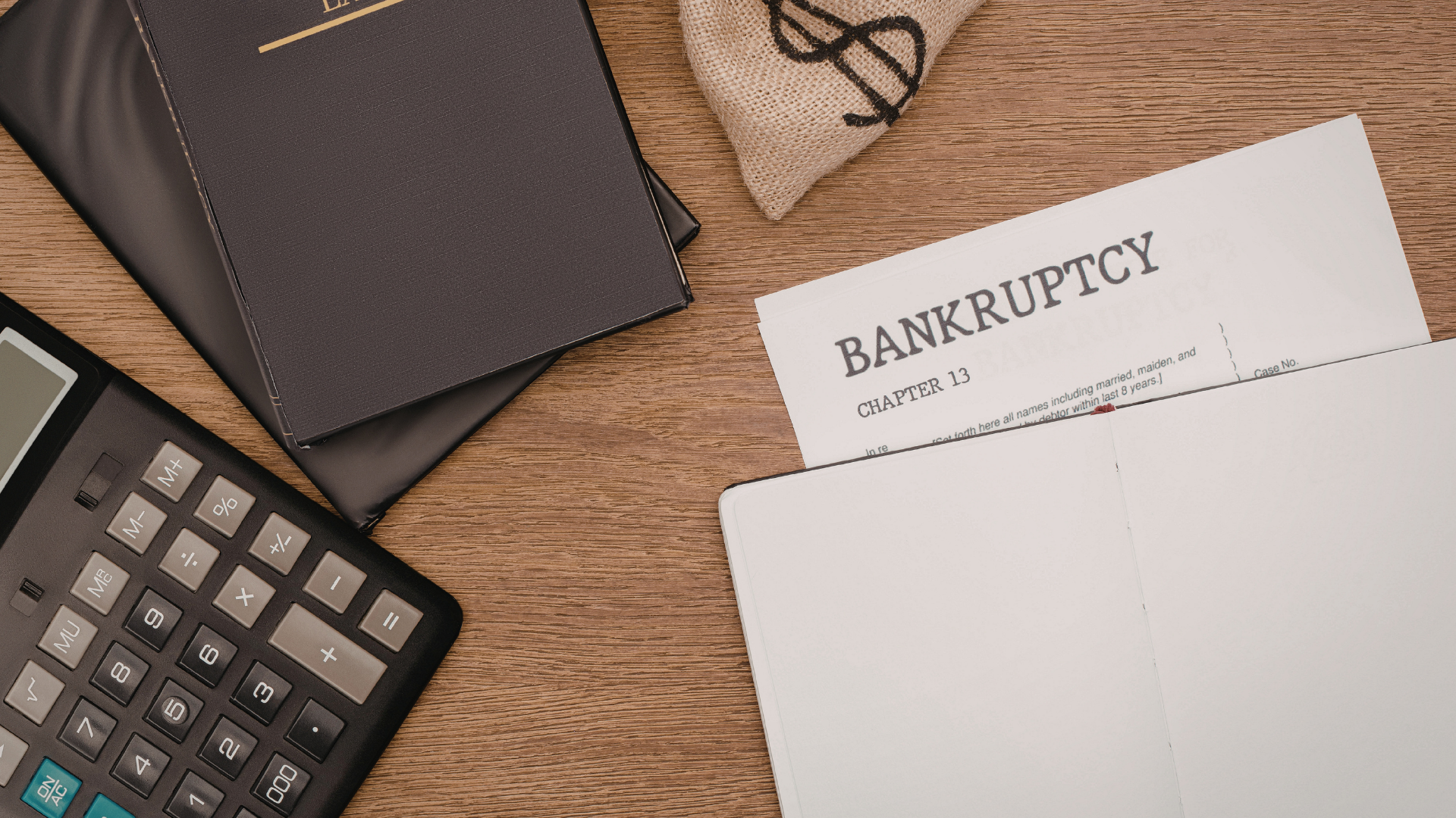 A top-down view of a calculator, a dark notebook, a money bag, and a bankruptcy form on a wooden table.