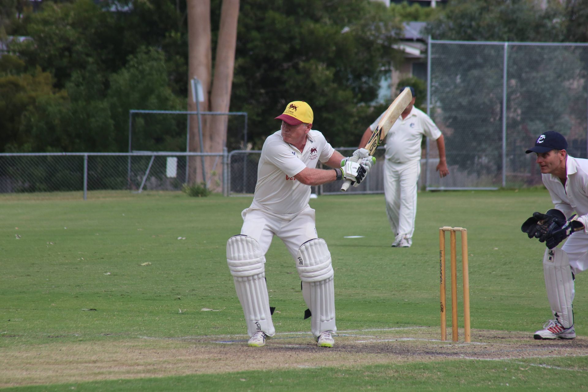Cricket batsman in white and yellow swings at ball, with wicketkeeper and bowler on a grassy field.