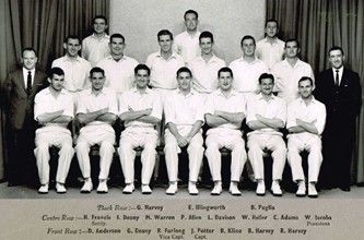 Black-and-white team portrait of 18 men in white uniforms, seated and standing in rows with name labels below