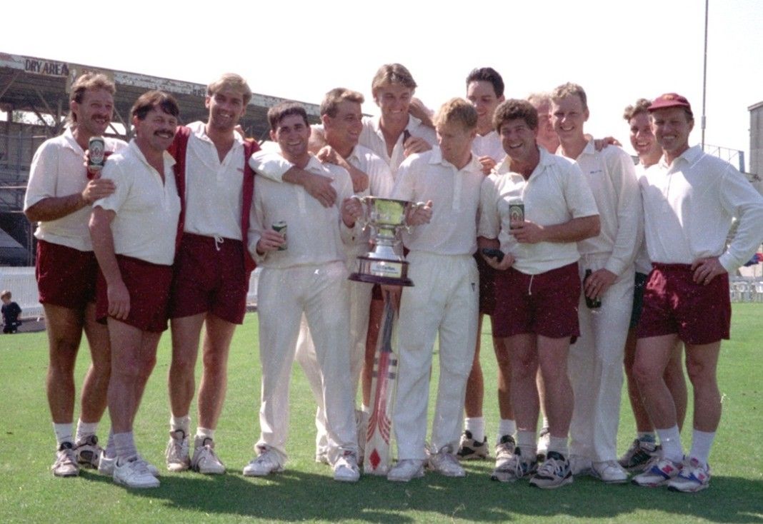 Cricket team in white and maroon uniforms posing on a field with a trophy at center