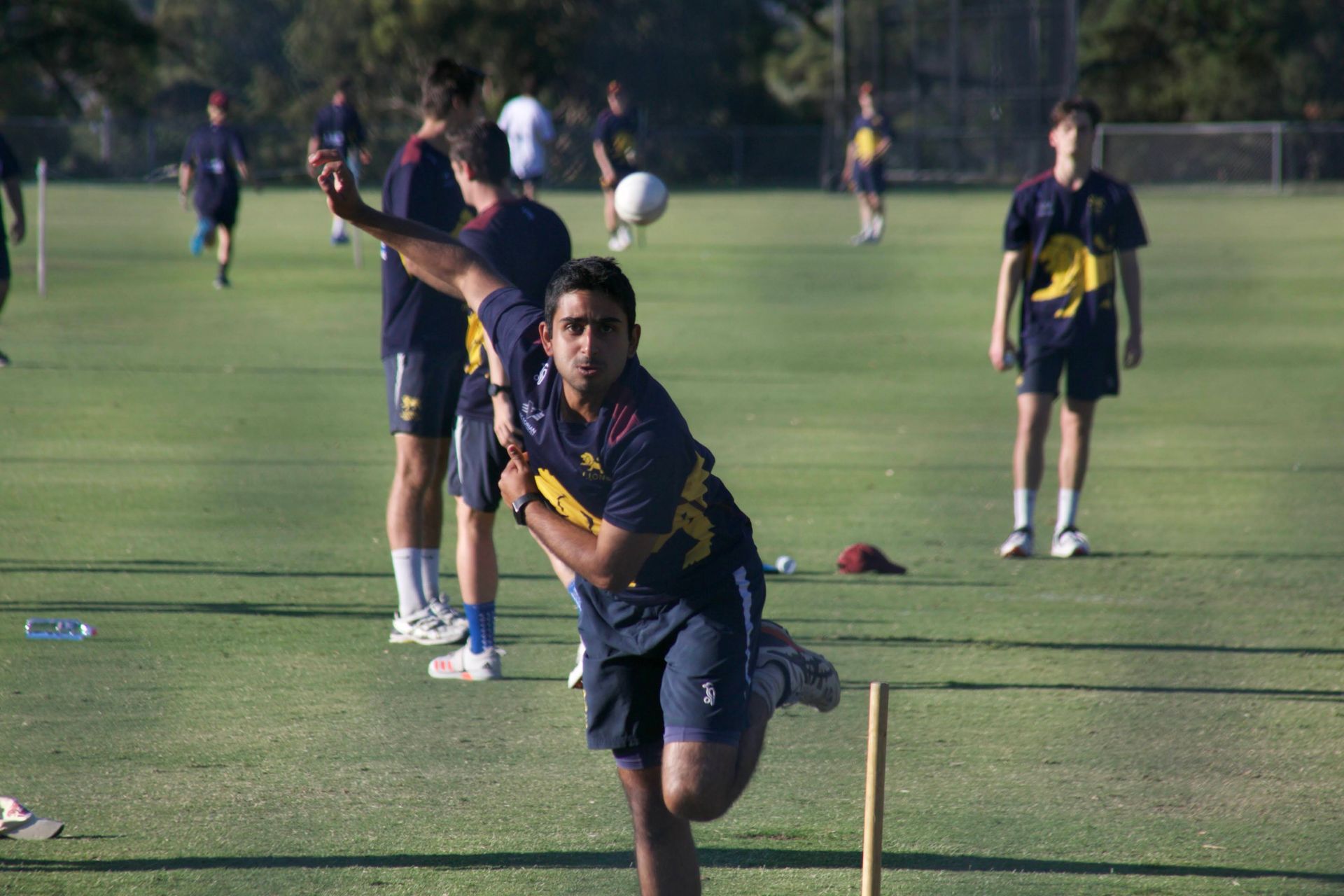 People practicing cricket on a grassy field, with one player bowling in the foreground