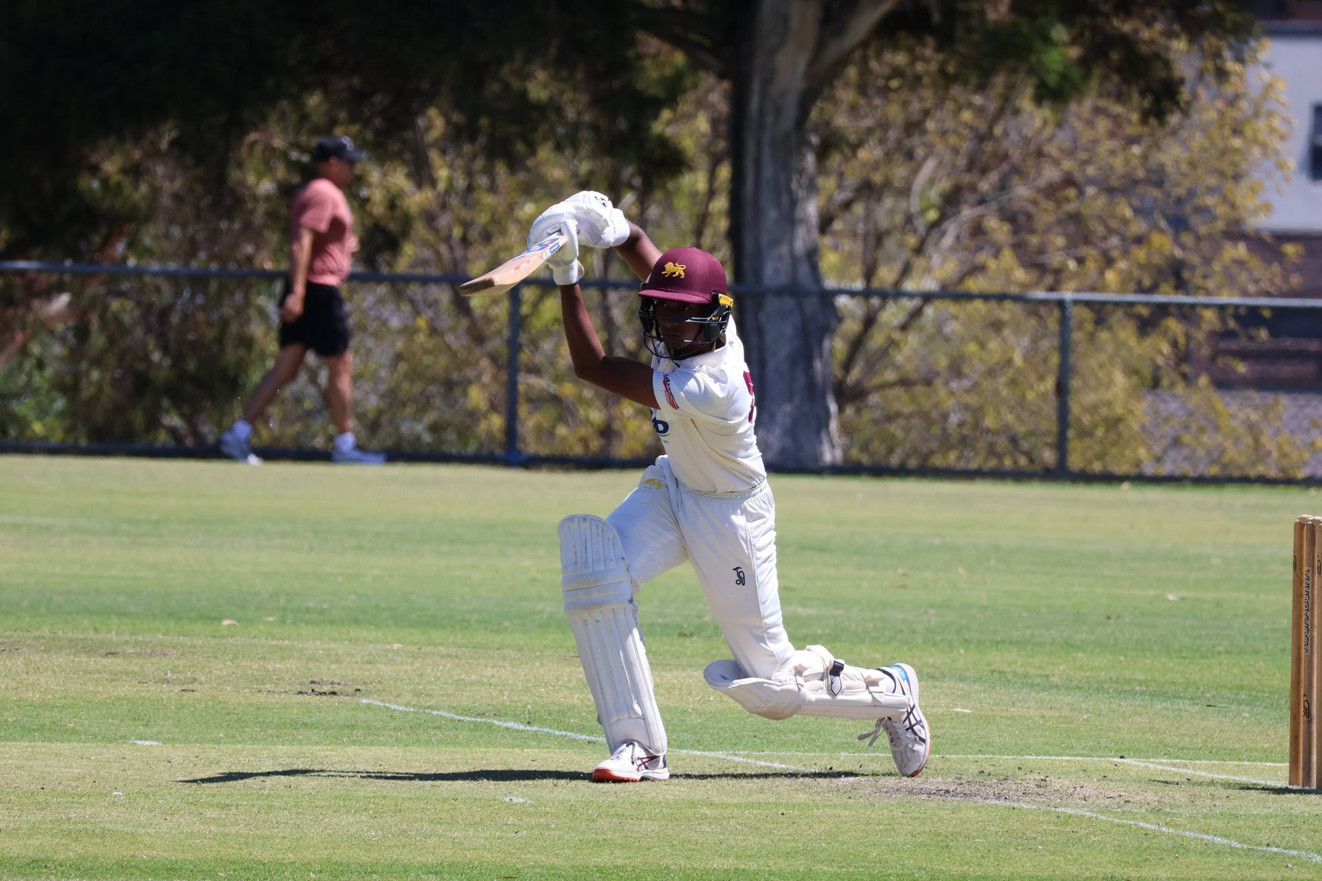 Cricketer in white swings at a ball on a grassy field, with a fielder in the background.