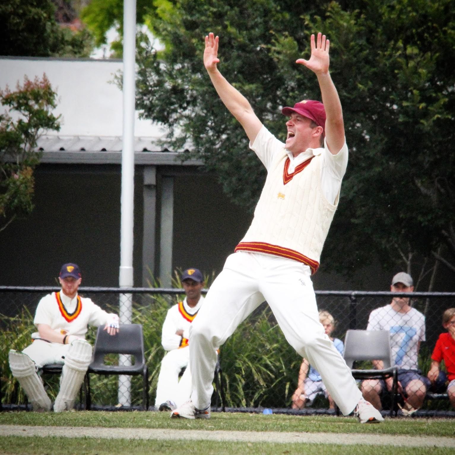 Cricket player in white uniform and red cap appeals with both arms raised on a field.
