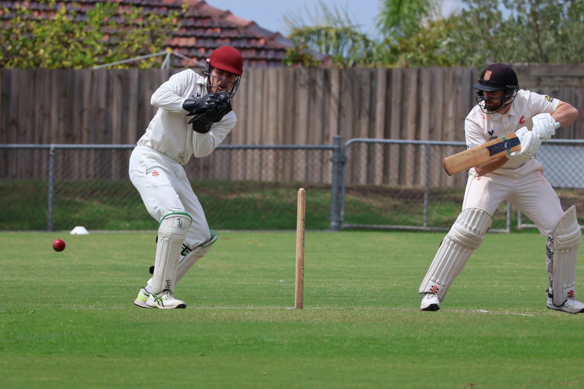 Two cricket players in white uniforms on a grassy field, one batting and one fielding near the stumps.