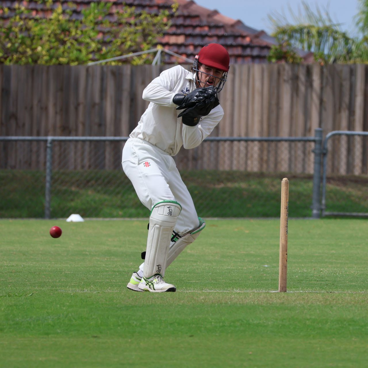 Cricket player in white gear and red helmet catches a red ball near the wicket on a grassy field