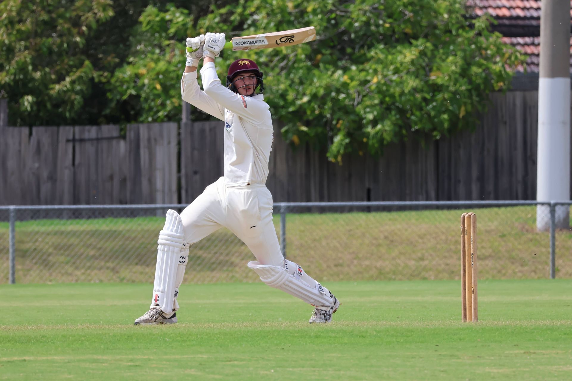 Cricketer in white mid-swing on a grassy field, wicket visible behind him.