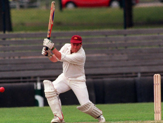 Cricket batsman in white uniform and red helmet swings at a ball on a field
