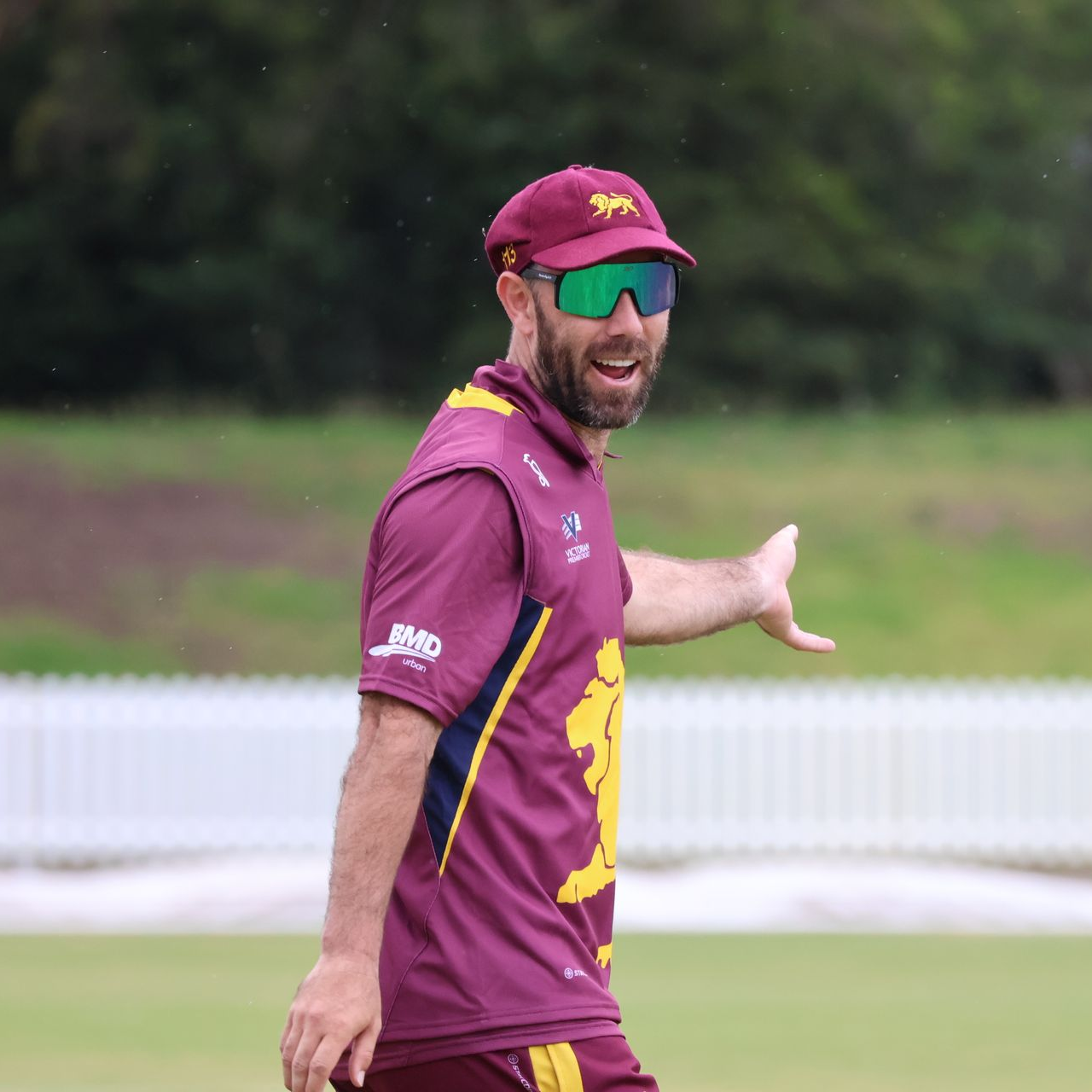 Cricketer in maroon and yellow uniform gesturing on a grassy field, wearing sunglasses and cap.