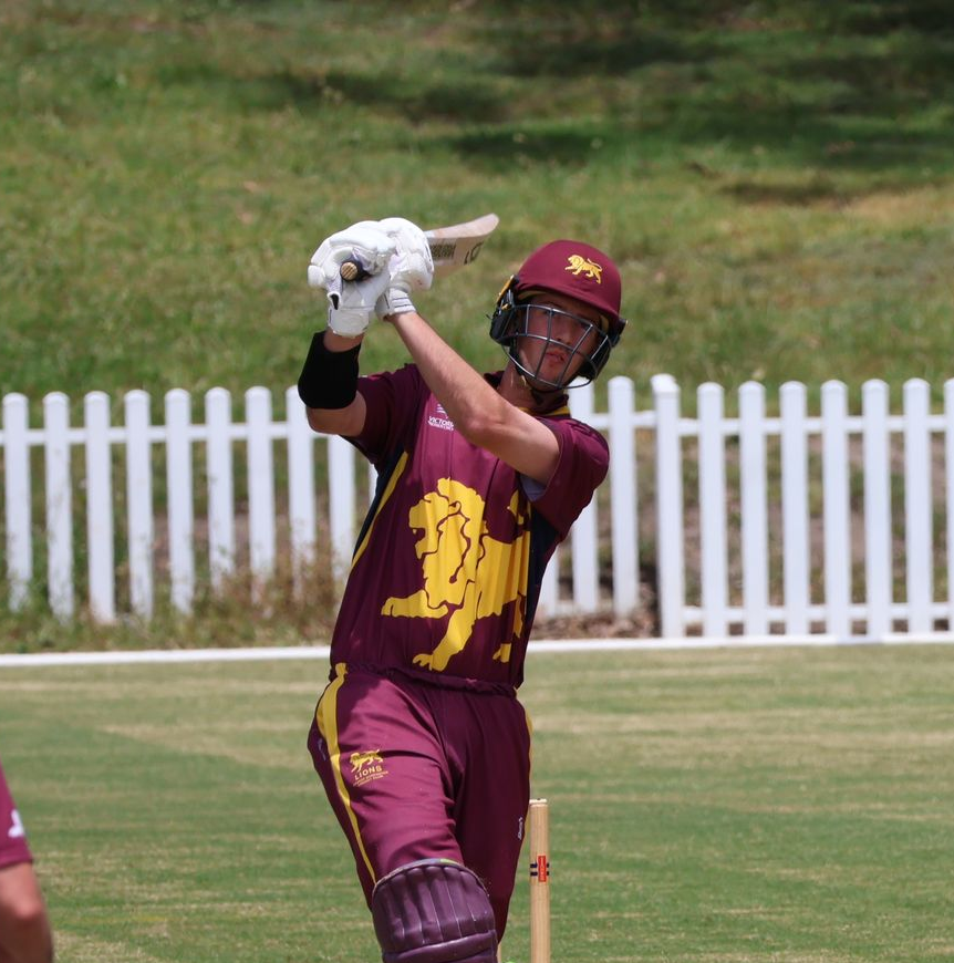 Cricketer in maroon and gold uniform swings a bat on a grassy field, with white fence in the background