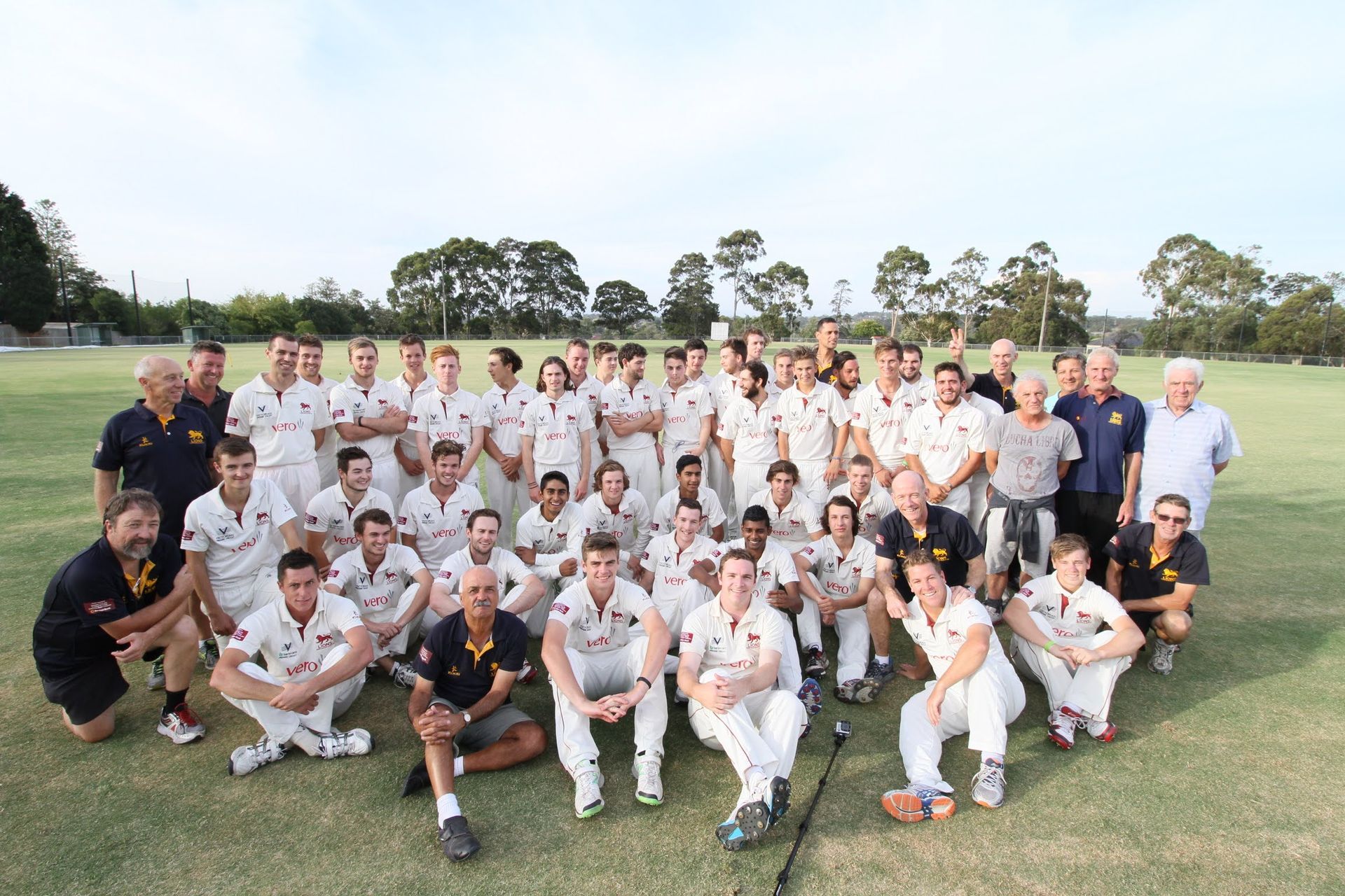 Cricket team group photo in white uniforms on a grassy field with coaches standing behind them