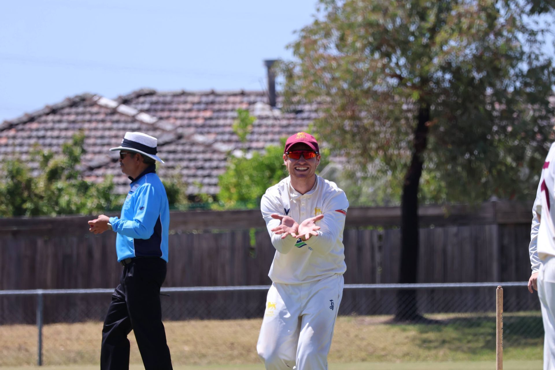 Two people in cricket uniforms on a grassy field, one in white and red reaching out, one in blue standing nearby.