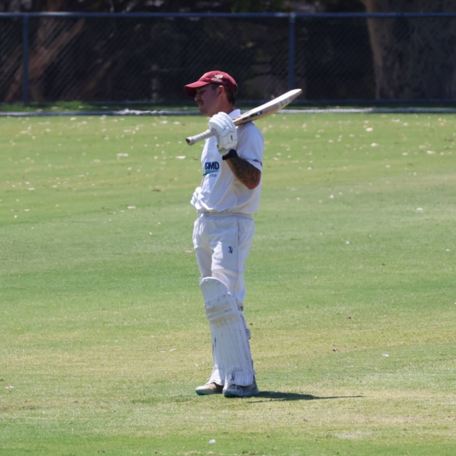 Cricket player in white uniform and red cap standing on a grassy field with a bat over his shoulder