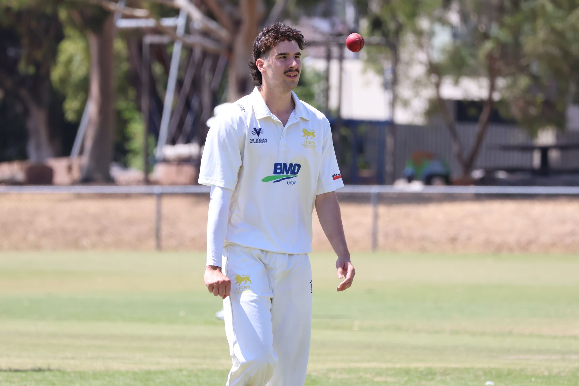 Cricketer in white uniform walking on a grassy field with a red ball in the air nearby