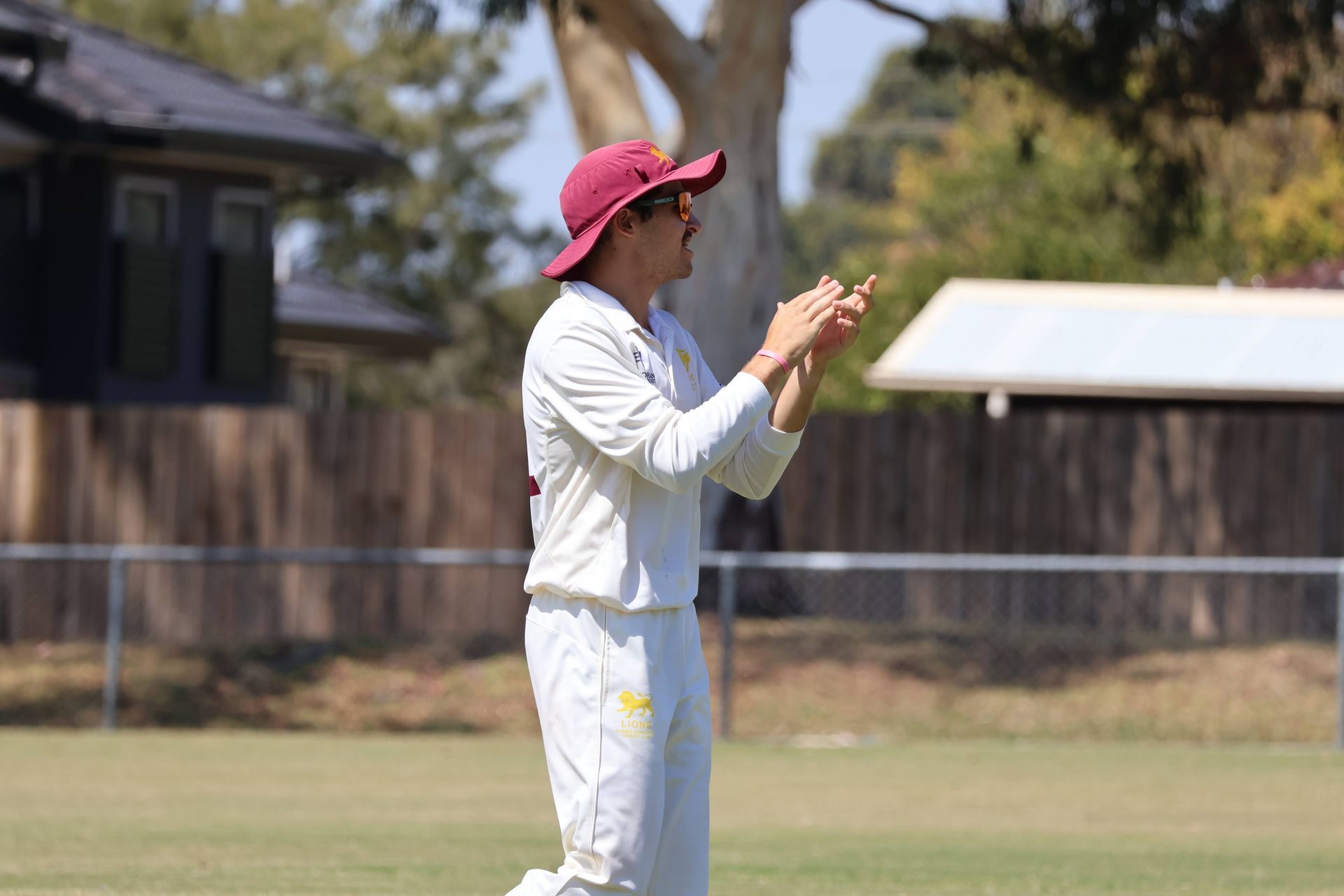 Cricketer in white uniform and pink hat clapping on a grassy field with trees and a fence in the background