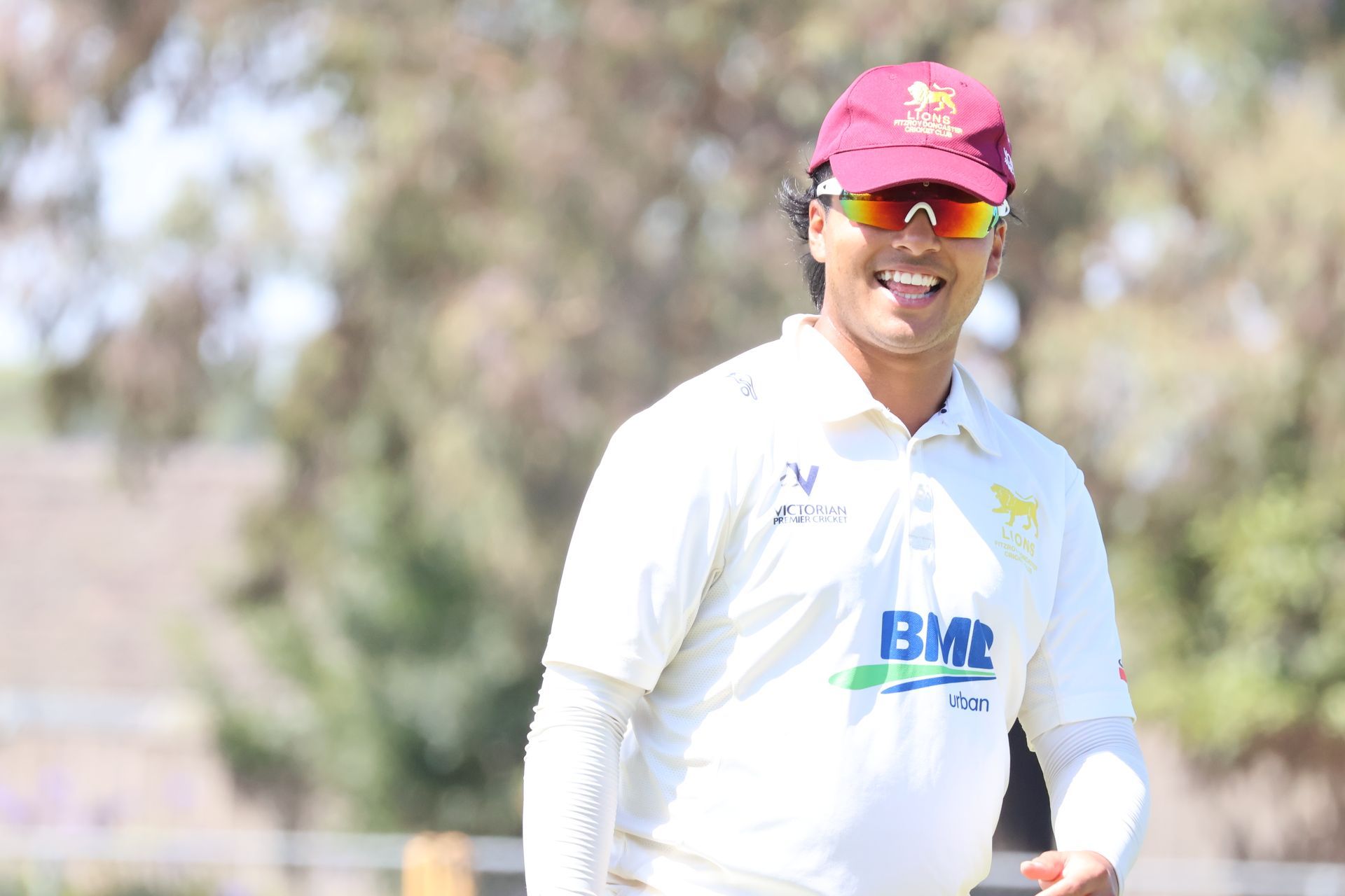 Cricketer in white uniform and maroon cap smiling on a sunny field