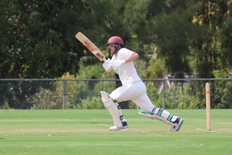 Cricket batter in white uniform runs on a grassy field, swinging a bat near the wicket.