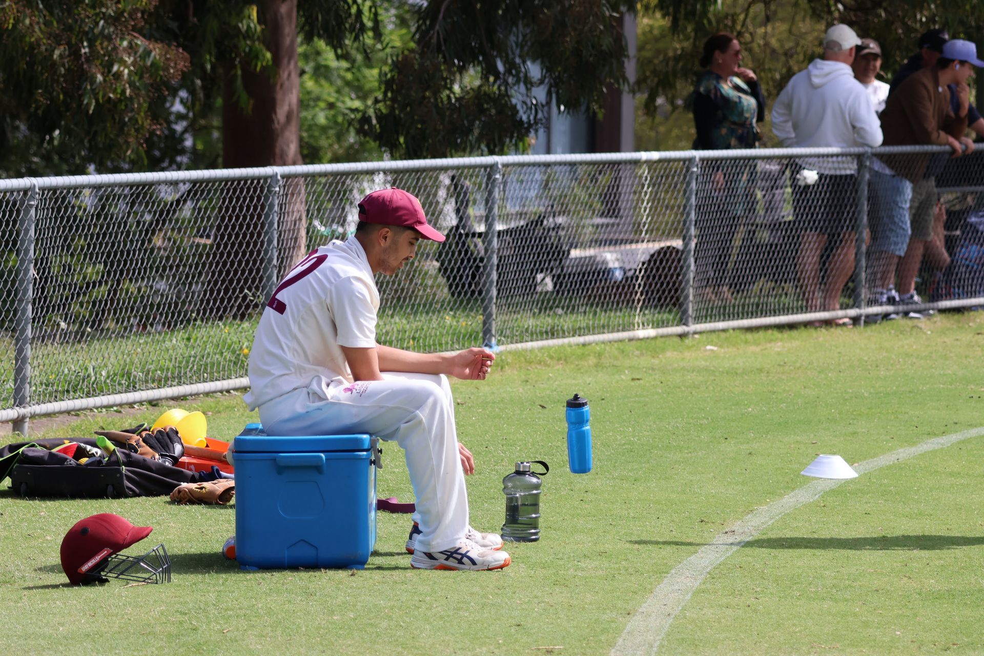 Person in white uniform and red cap sits on a blue cooler by a field fence, holding a drink bottle.