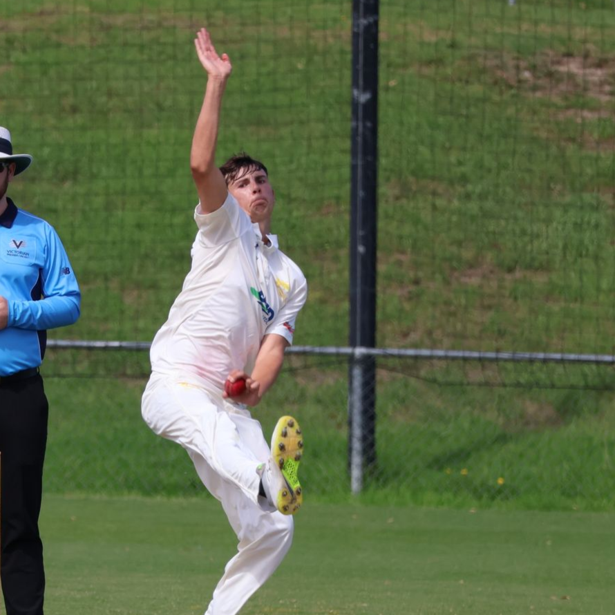 Cricketer in white uniform mid-bowl, arm raised, on a green pitch with an umpire nearby.