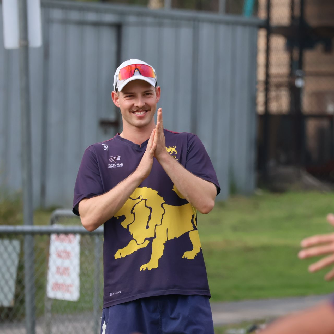 Smiling person in a navy shirt with a yellow lion graphic, clapping outdoors near a fence