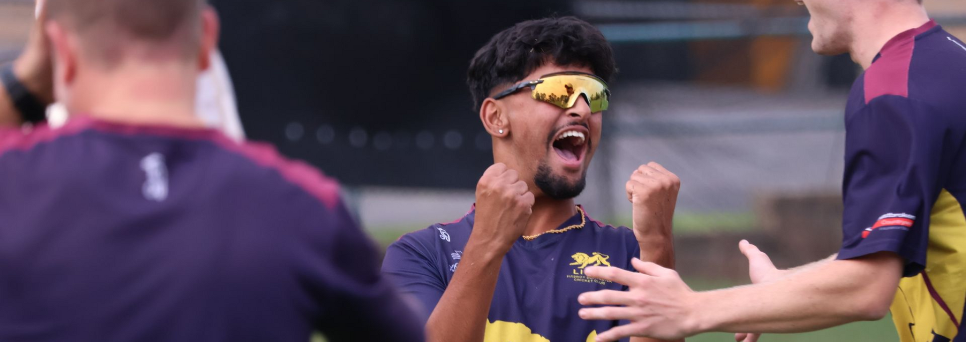 Cricket players in navy and yellow celebrate on the field, one shouting with raised fist.