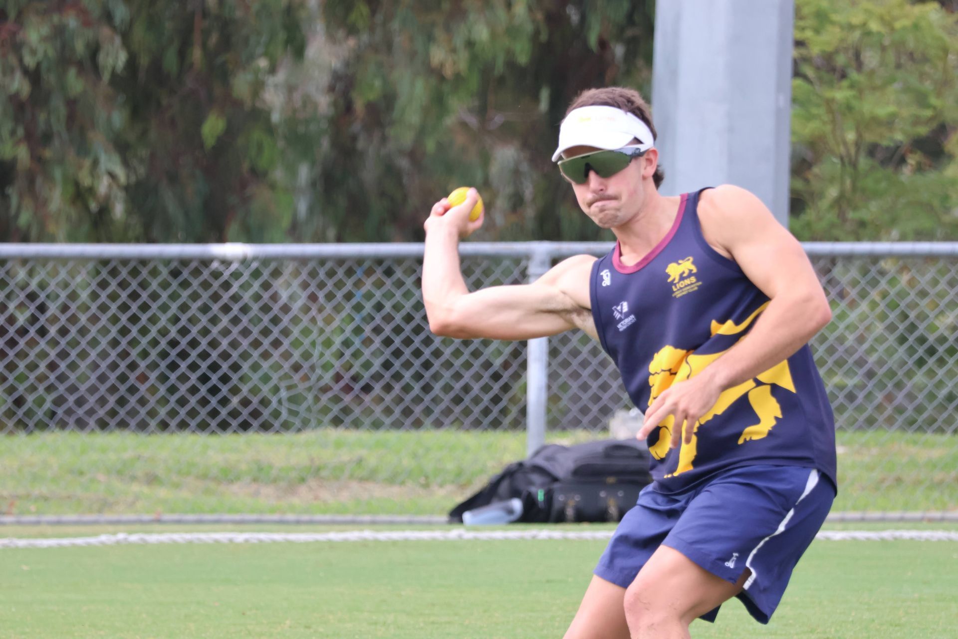 Athlete in navy and yellow uniform throws a ball on a grassy field, wearing a visor and sunglasses.