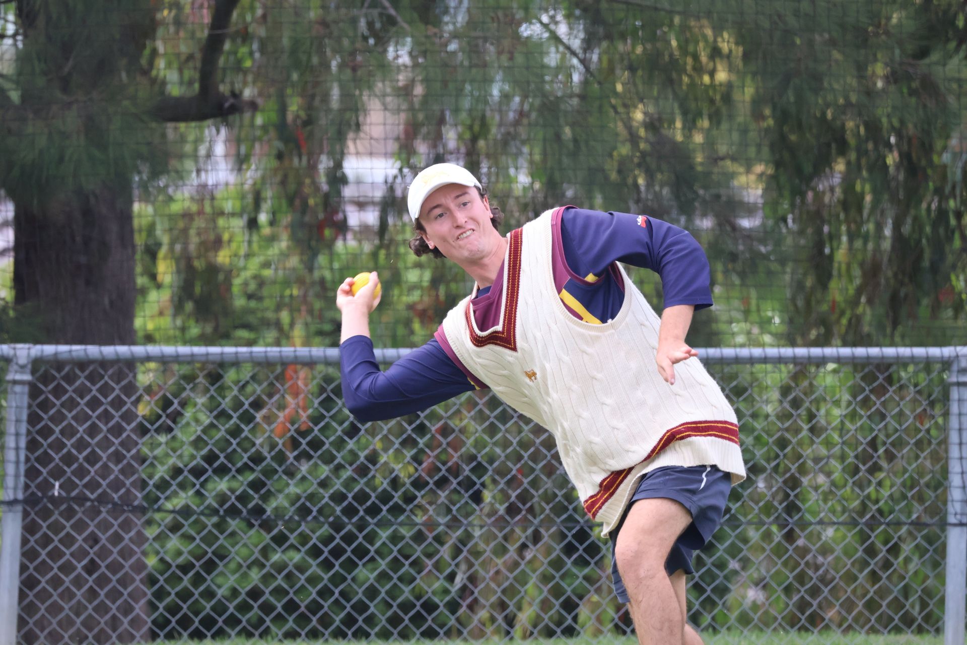 Man in a white cap and vest throwing a ball on a field near a chain-link fence