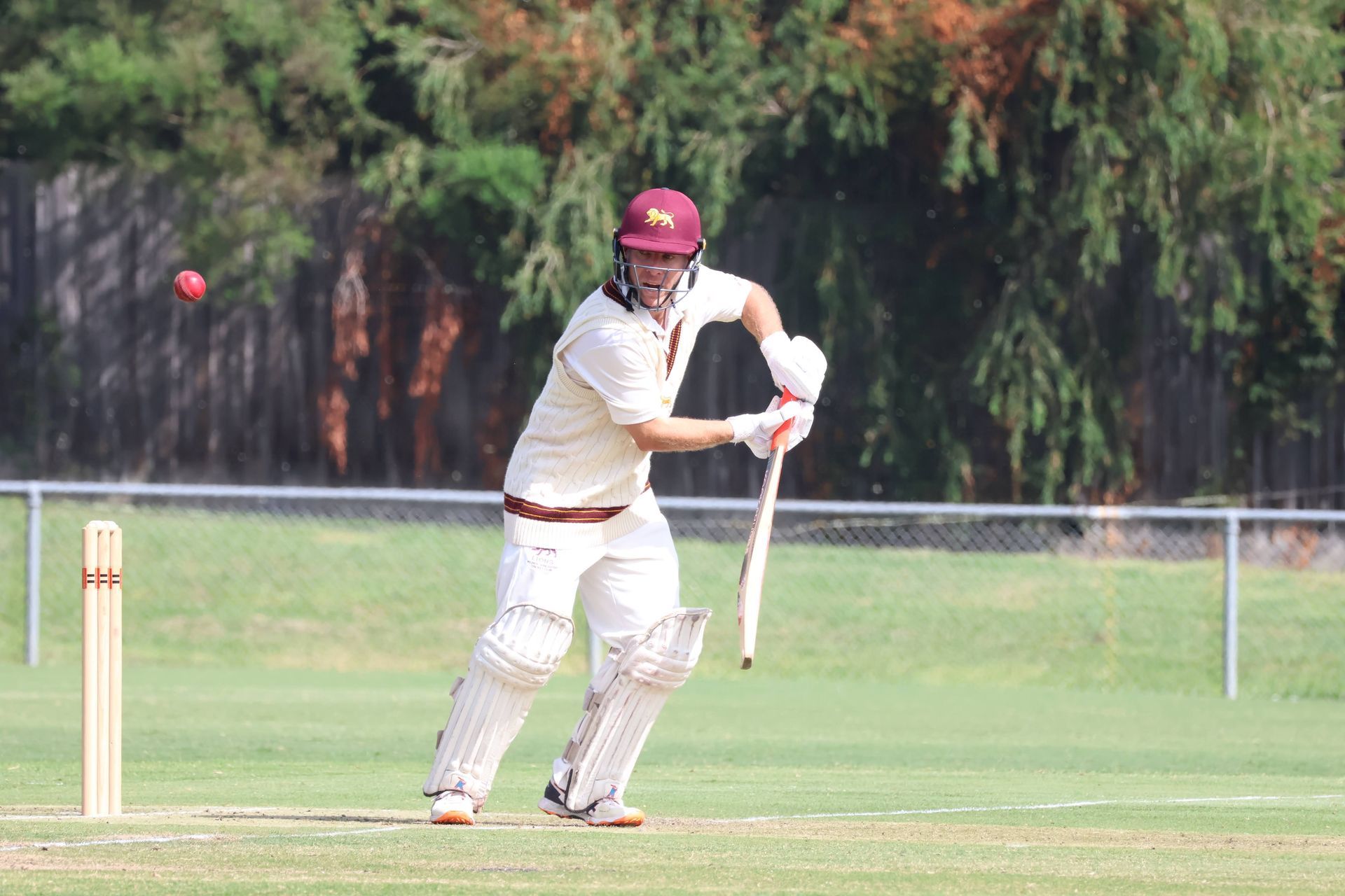 Cricket batter in white uniform and maroon helmet, poised with bat near the wicket on a grassy field.
