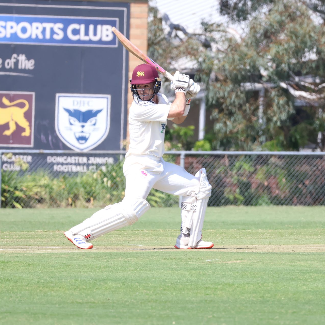 Cricket batter in white kit mid-swing on a grassy field, with sports club signs in the background