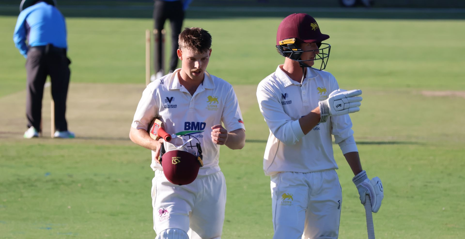 Two cricket players in white uniforms walking on a field, one holding a helmet and the other reading a card.