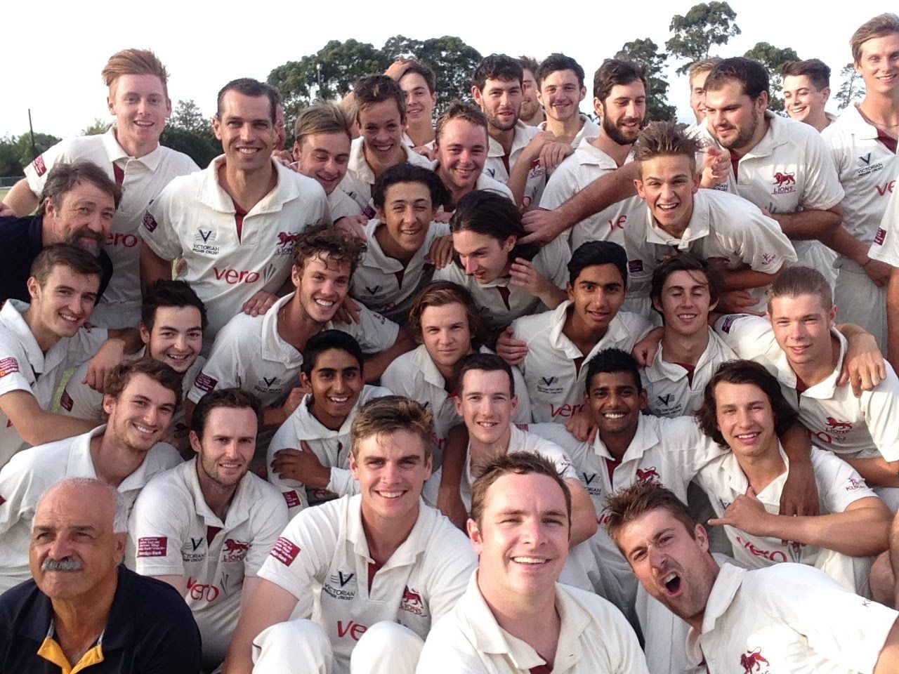 Large group of people in white sports uniforms posing closely together outdoors, smiling at the camera.