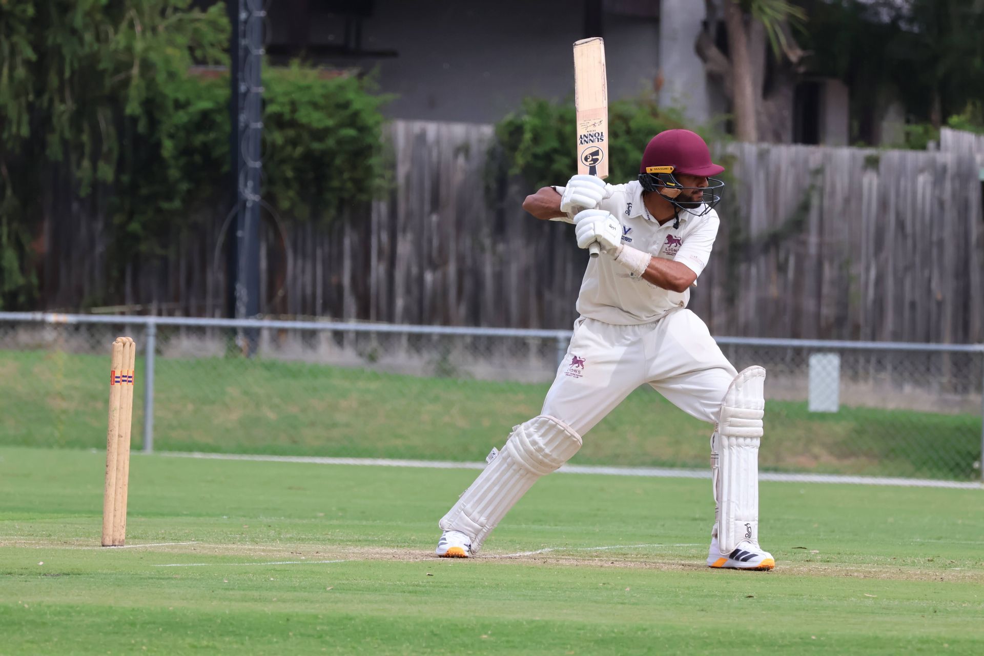 Cricket batter in white uniform and maroon helmet playing a shot on a grassy field