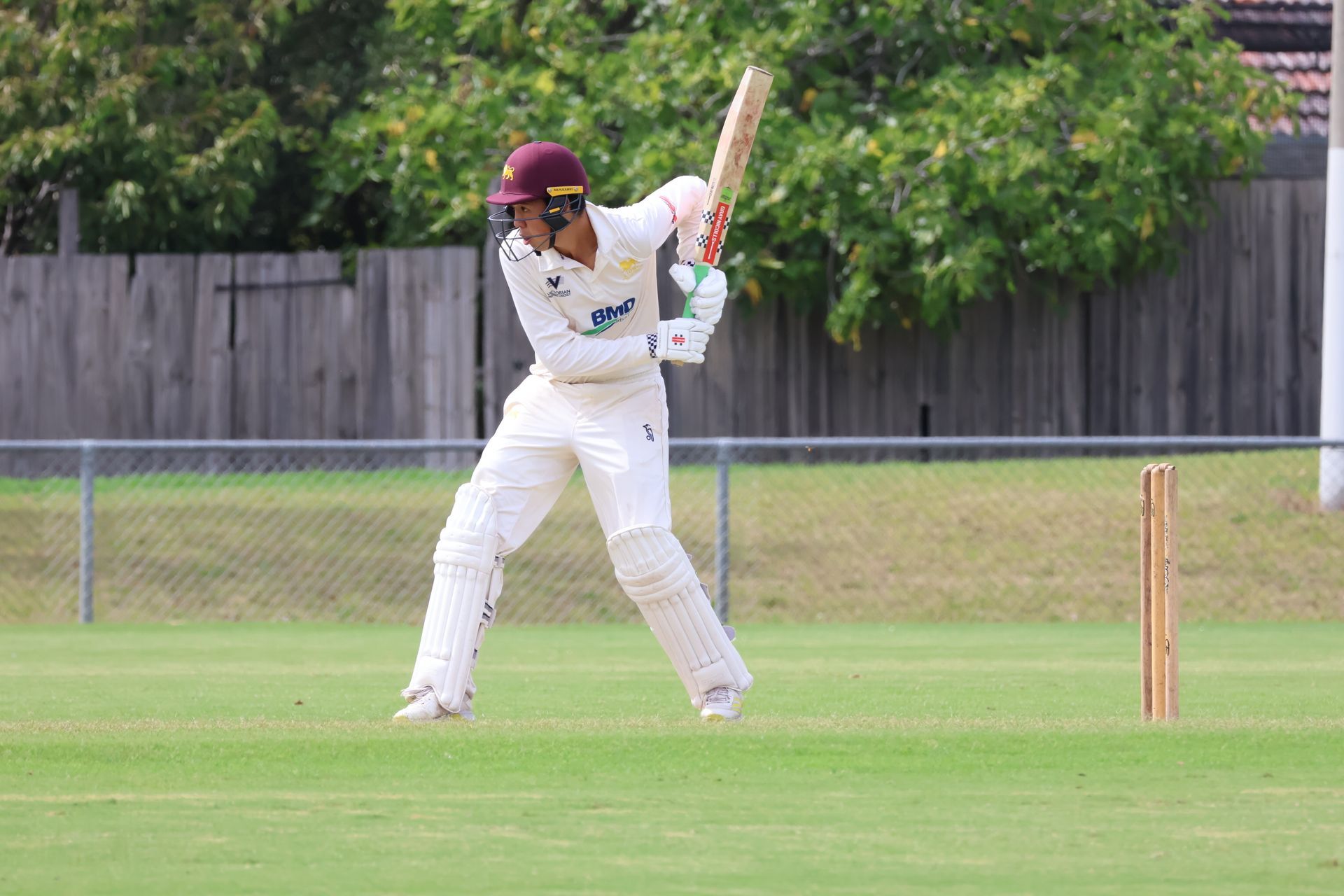 Cricketer in white batting gear plays a shot on a grassy field, with stumps behind him.