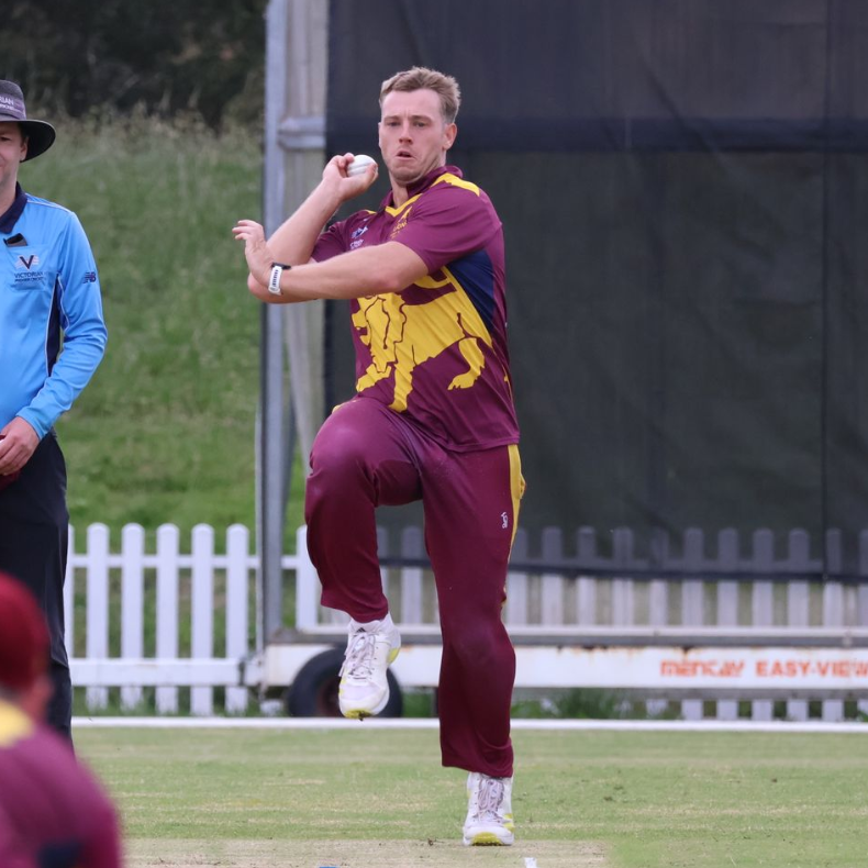 Cricketer in maroon and gold uniform mid-bowling action on a grassy field