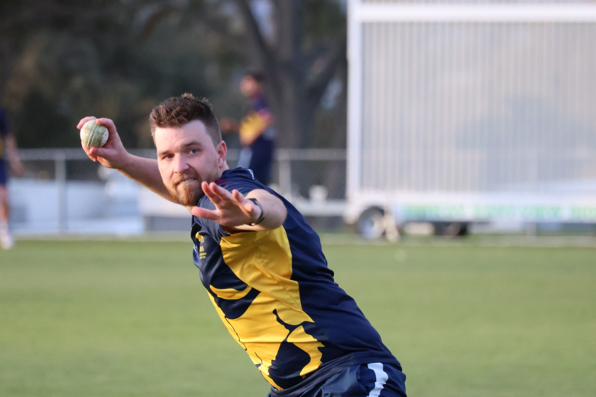 Cricketer in blue and yellow uniform throwing a ball on a grassy field