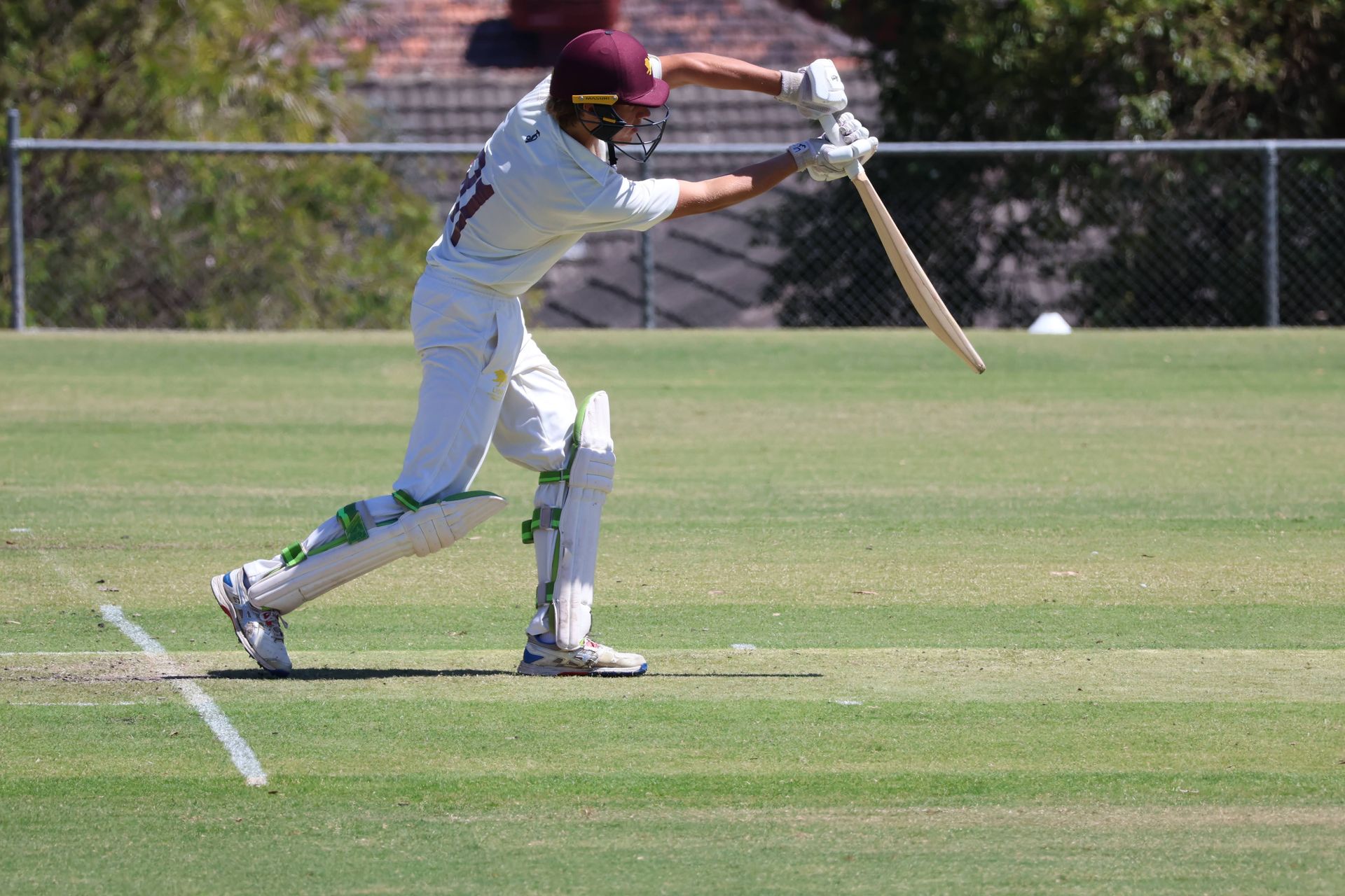 Cricketer in white batting on a sunny field, swinging at a ball with wicket visible behind.
