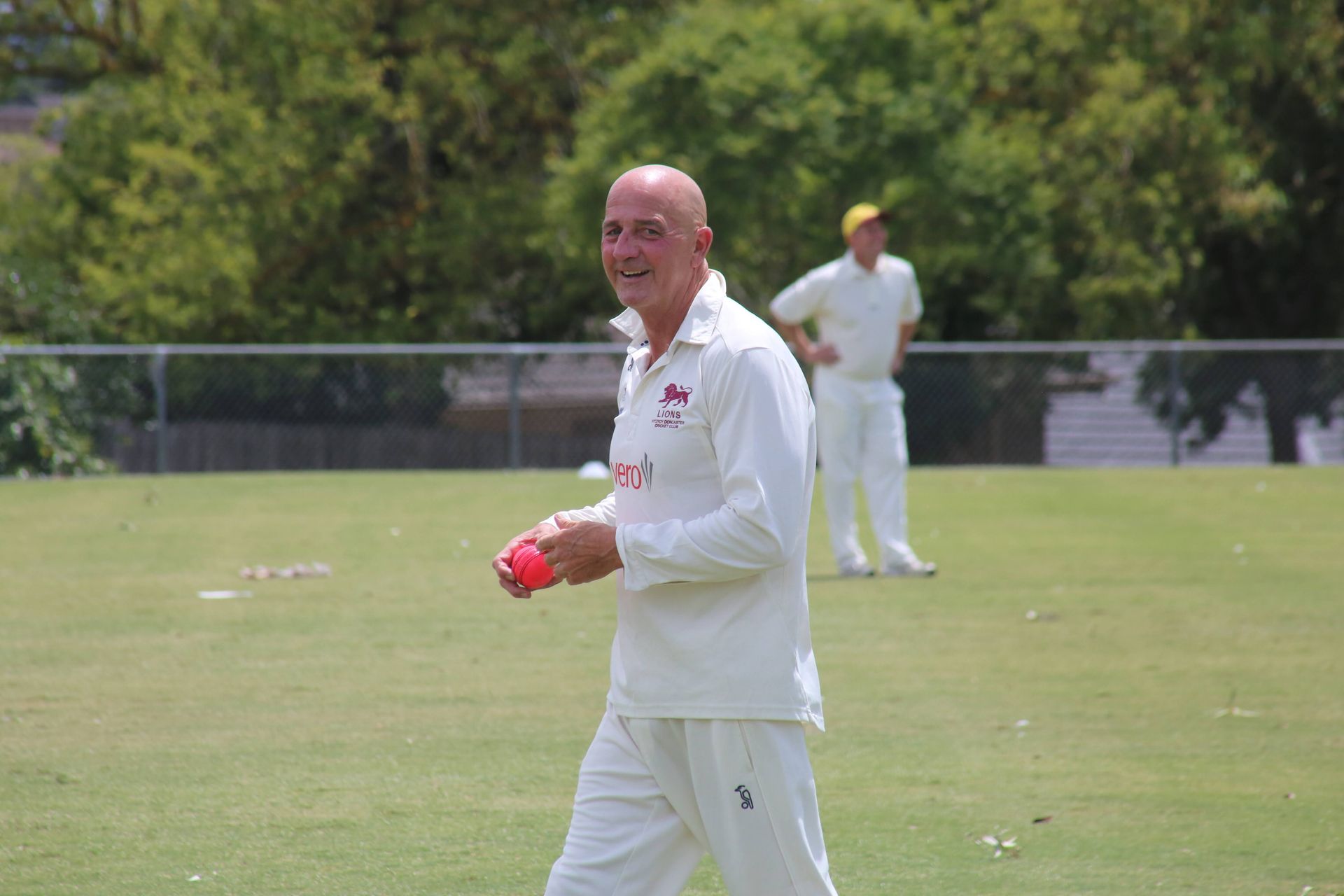 Cricket player in white uniform holding a red ball on a grassy field, with another player in the background