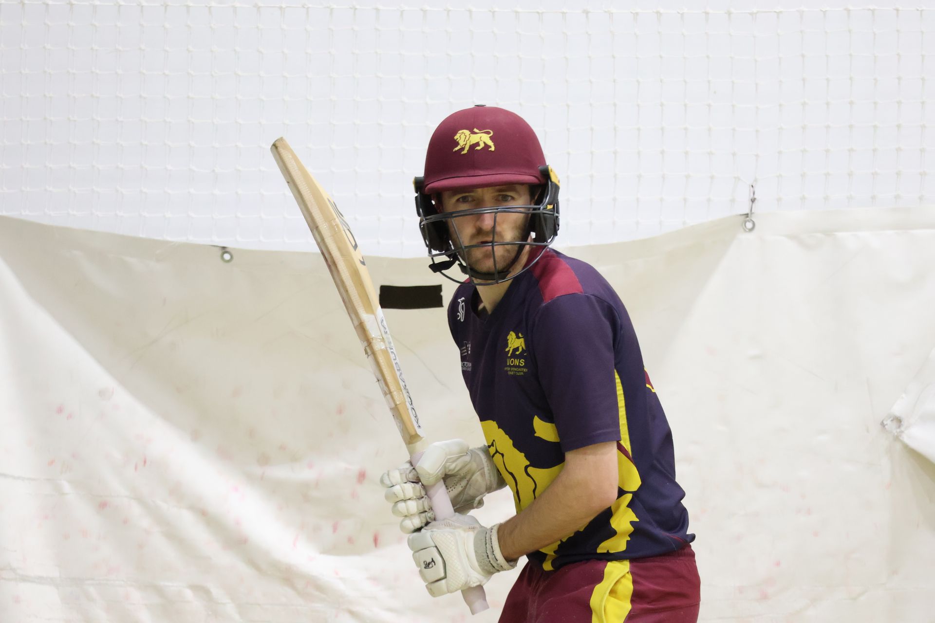 Cricketer in maroon and gold uniform holding a bat in front of a white practice net